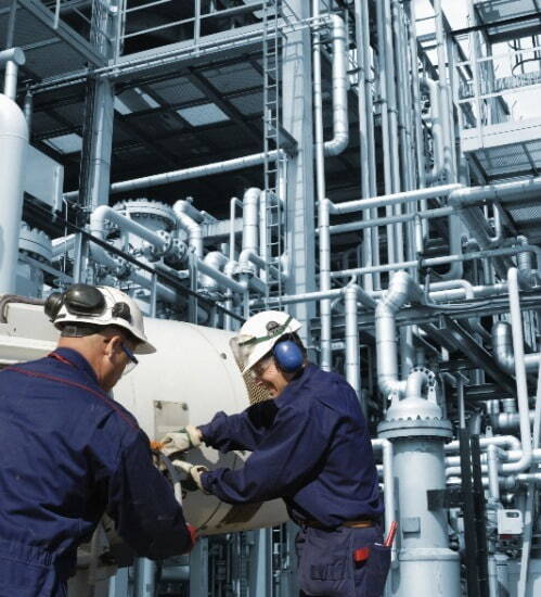 2 men in blue overalls wearing white hard hats and ear defenders, working on equipment 